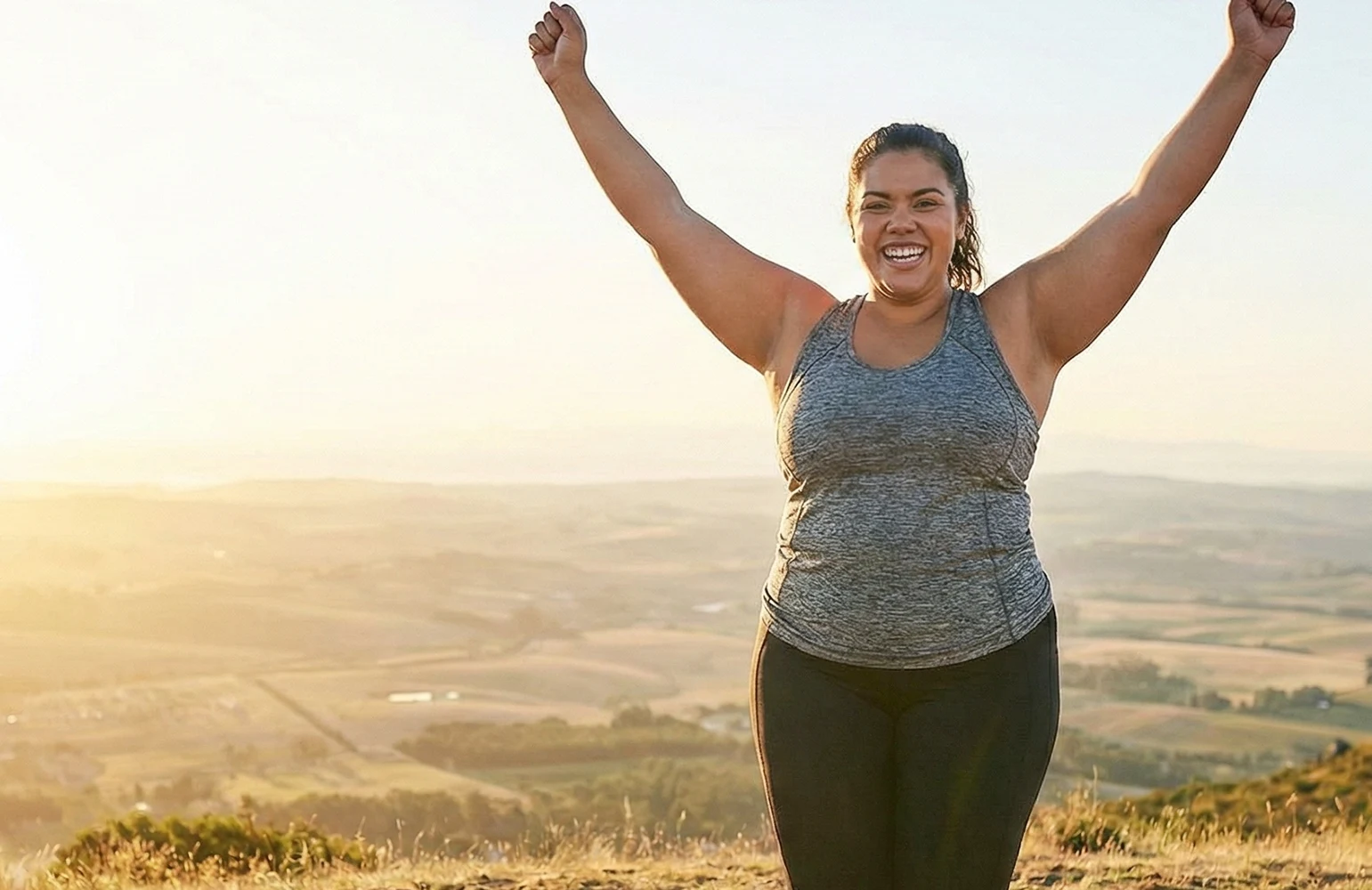 Abnehmspritze mit ärztlicher Begleitung im CUROS urologischen Zentrum Köln Bonn - Hero4-mobi Das Symbolbild zeigt eine übergewichtige Frau, die in Sportkleidung auf der Spitze eines Hügels steht und glücklich die Arme in die Luft reckt. Abnehmen mit der Abnehmspritze unter ärztlicher Betreuung bei CUROS
